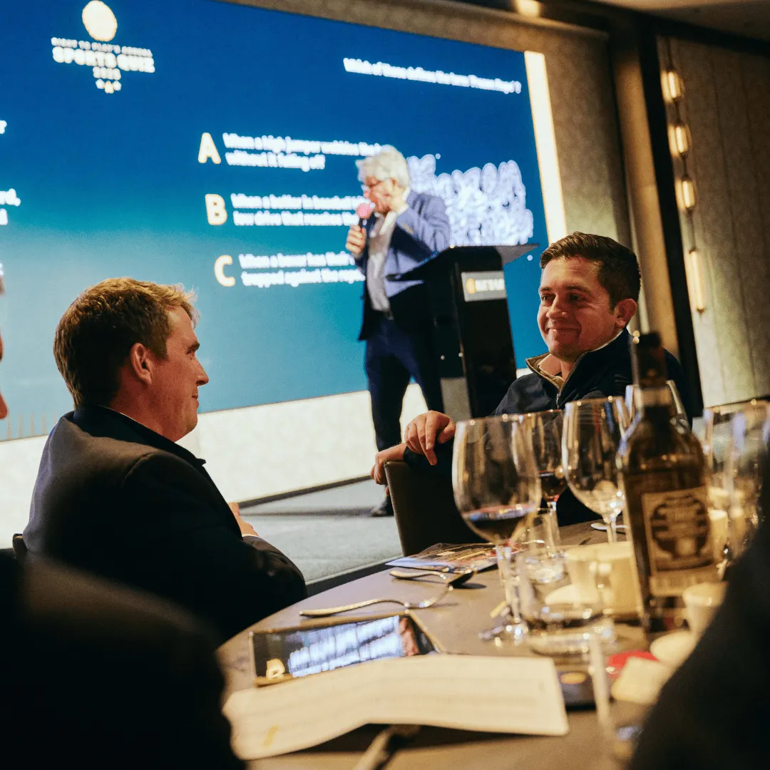 Guests smile at each other on a table in front of the stage, with John Inverdale running the quiz in the background