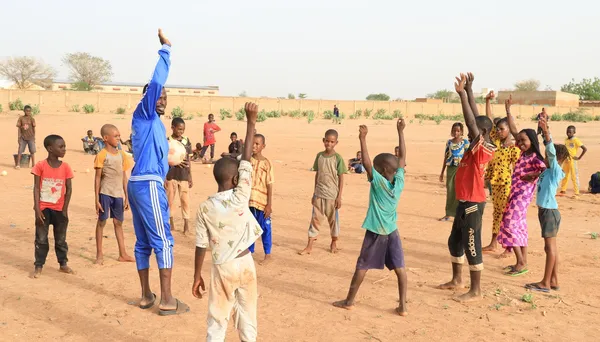 A group of children are engaged in an outdoor activity, with one individual in a blue tracksuit leading them, while others raise their arms in unison.