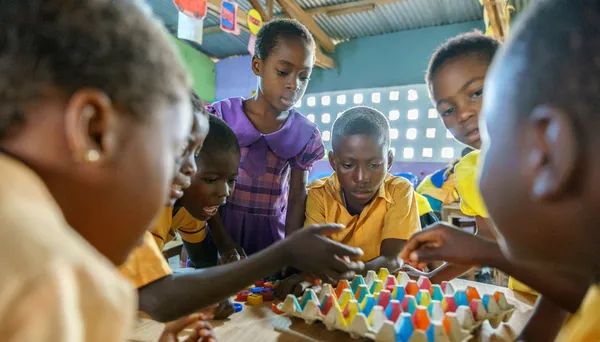 A group of children are gathered around a table, looking down at colorful objects they are arranging.