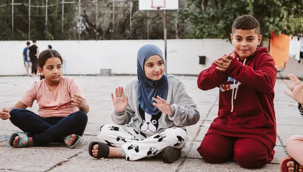 Three children sitting on the ground, two are wearing headscarves, and one is wearing a red hoodie, all gesturing with their hands.