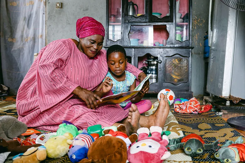 A person in a pink outfit sits on the floor with a child, reading a book surrounded by toys.